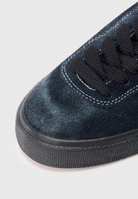 Close-up de um dedo de ténis em suede azul escuro com atacadores pretos e sola de borracha texturizada preta em fundo branco.