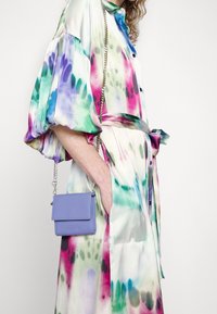 Femme portant une robe tie-dye colorée avec des manches bouffantes et une taille nouée, portant un petit sac bandoulière lavande avec une chaîne argentée.
