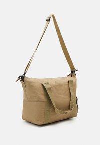 Khaki-kangaslaukku, jossa on kaksinkertaiset yläkahvat, irrotettava olkahihna ja mustat vetoketjeyksityiskohdat. Ominaisuuksiin kuuluu rakenteellinen suorakulmainen muoto.