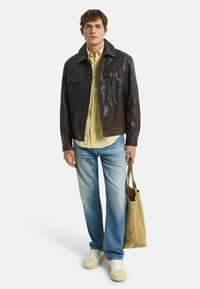 Dunkelbraune Lederjacke mit zwei Brusttaschen, hellgelbes Hemd, ausgewaschene blaue Jeans und wei�ße Sneaker. Hält eine braune Stofftragetasche.