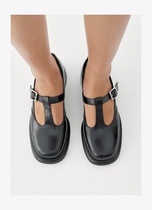 Čierne kožené topánky s dizajnom T-remenčeka, uzavretou špičkou a striebornými prackami. Hladký povrch s zaoblenou špičkou a plochou podrážkou.