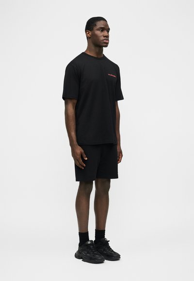 Homme debout portant un T-shirt noir avec un petit texte rouge, un short noir, des chaussettes noires et des baskets noires sur un fond clair uni.