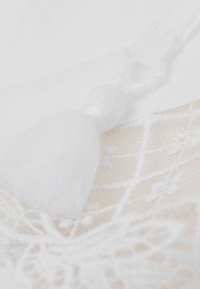 Tela blanca con un intrincado patrón de encaje, que presenta un acento de borlas blancas. La textura es suave y delicada, destacando los finos detalles.
