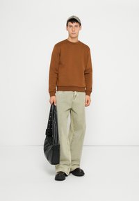 Hnedý sveter s okrúhlym výstrihom, svetlozelené voľné džínsy a čierne slip-on topánky. Drží čiernu, textúrovanú kabelku cez rameno s cvočkovým popruhom.