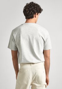 Camiseta de algodón gris claro con mangas cortas, escote redondo y textura suave, combinada con pantalones beige que tienen dos bolsillos traseros.