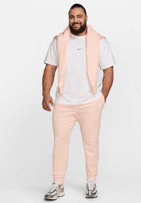 Sudadera de Nike en rosa blush caída sobre los hombros, combinada con una camiseta blanca y pantalones de chándal en rosa blush a juego. Incluye sutiles acentos del logo.