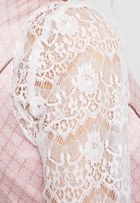 Manche en dentelle blanche avec des motifs floraux complexes, associée à un fond texturé en quilt rose. Présente un tissu transparent et des détails délicats.