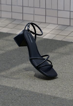 Black heeled sandal with an open toe, featuring thin strap design and a square heel. Smooth texture with a glossy finish. Brand printed inside.