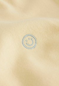 Tela de algodón color crema con una textura suave que presenta un logo de cara sonriente bordado en azul pálido en el centro.