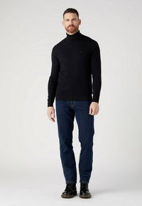 Czarny sweter z golfem, dopasowany krój, prążkowane mankiety i dół, noszony z ciemnoniebieskimi dżinsami i czarnymi sznurowanymi butami. Model stoi w pozycji wyprostowanej, na neutralnym tle.