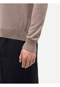 Beige stickad tröja med figursydd design, ribbade muddar och fåll, bärs över mörka byxor. Synlig textur och sömlös finish i mjukt tyg.