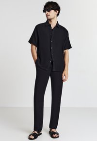 Schwarzes kurzärmliges Hemd mit Knöpfen, aus strukturiertem Stoff, kombiniert mit schwarzen Slim-Fit-Hosen und schwarzen Slippern, getragen mit dunklen Sonnenbrillen.