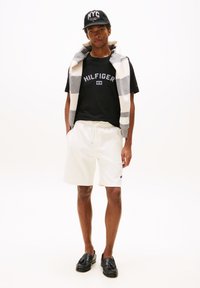 T-shirt en coton noir avec le logo "HILFIGER", shorts crème, pull rayé gris et blanc drapé, casquette noire avec "NYC", mocassins noirs.