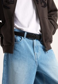 Ruskean vetoketjuhuppari brodeeratulla kuvalla, valkoinen crewneck-paita alla, vaaleansiniset farkut, tekstuurilla varustettu musta vyö logo-soljella.