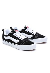 Paire de baskets basses noir et blanc avec lacets blancs, bande latérale blanche et logo rouge sur la languette, présentée sur fond blanc.