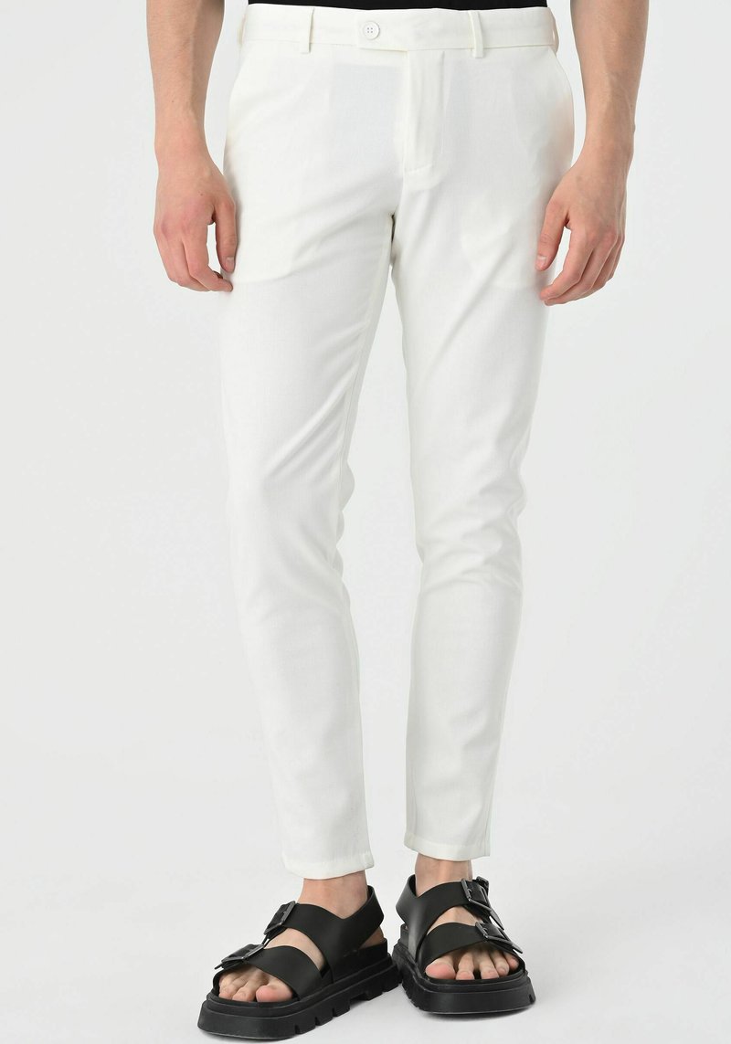 Man wearing slim-fit white trousers and black chunky sandals with buckle straps, standing on a plain light background.