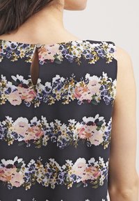 Donna che indossa un vestito nero senza maniche con strisce floreali orizzontali in rosa, viola, bianco e verde, scollo posteriore con apertura a chiave abbottonata.
