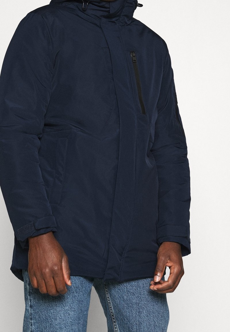Mand iført en marineblå polstret jakke med lynlåslomme på brystet og blå jeans, stående mod en ensfarvet lys baggrund.