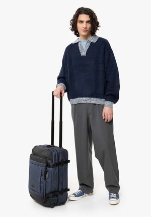 Navy ribbed sweater with a contrasting collar, gray pants, and blue sneakers beside a rugged blue and black wheeled suitcase with straps.