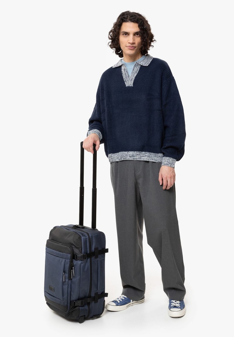 Suéter de canalé azul marino con un cuello en contraste, pantalones grises y zapatillas deportivas azules junto a una resistente maleta de ruedas azul y negra con correas.