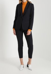 Marinblå skräddarsydd blazer, figurnära svarta byxor och en orange ribbad polotröja. Vita klackskor med blockklack fulländar looken. Släta texturer.