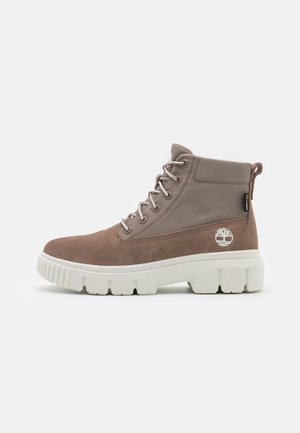 Beżowo-taupe wysokie buty sznurowane z grubą białą podeszwą, detalami szwów, pętelką i logo Timberland po boku.