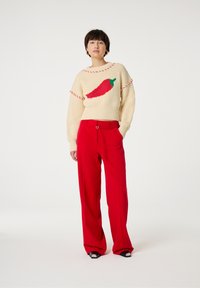 Creme kleurige trui met een ontwerp van rode chilipepers, gebreide textuur, gecombineerd met felrode broek met wijde pijpen en zwarte hakken met zilveren accenten.