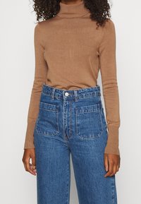 Brązowy sweter z golfem w zestawie z niebieskimi dżinsami z wysokim stanem. Sweter ma dopasowany krój; dżinsy mają szerokie nogawki i kieszenie z przodu.