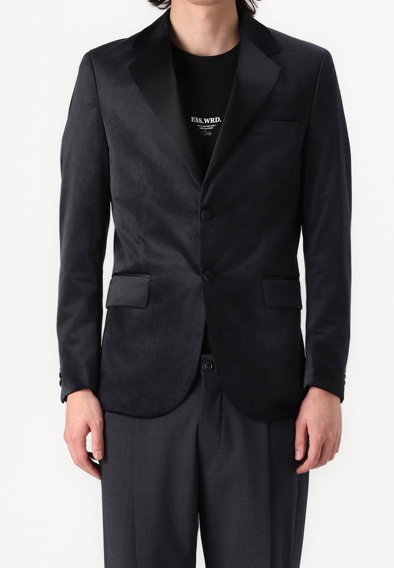 Svart blazer med satinklappor, två knappar och sidofickor. Slät yta, figursydd design, bärs över en svart T-shirt.