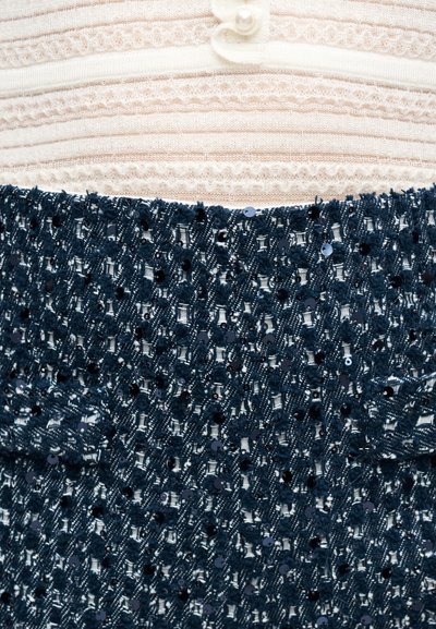 Άσπρο πλεκτό ύφασμα με μαργαριταρένιο κουμπί πάνω από υφασμα με υφή από σκούρο μπλε παγιέτες με υφαδωτά σχέδια.