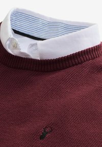 Pull en maille bordeaux superposé à une chemise blanche à boutons avec un col intérieur rayé bleu et un logo cerf brodé noir sur la poitrine.
