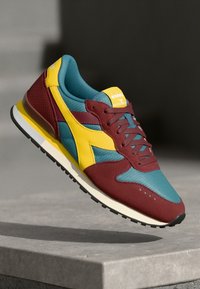 Běžecká obuv s maroon kůží a teal síťovinou na svršku, žlutými akcenty a černou gumovou podrážkou s texturovanými detaily.