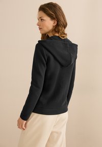 Sudadera negra hecha de una tela suave, con un diseño ajustado, bolsillo tipo canguro y capucha. La parte trasera muestra un acabado texturizado y detalles de costura.