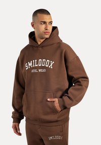 Le sweat à capuche marron présente une grande poche avant, un logo brodé blanc avec l'inscription "SMILODOX ATHL. WEAR," ainsi que des poignets et un ourlet côtelés.