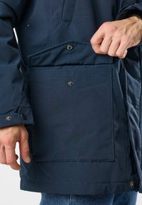 Man fastening snap button on large navy blue jacket pocket, showing detailed stitching and buttoned cuff over blue jeans.