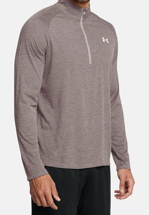 Homme portant un pull de sport mauve à manches longues avec une fermeture éclair quart de zip et logo blanc Under Armour sur le côté gauche de la poitrine.