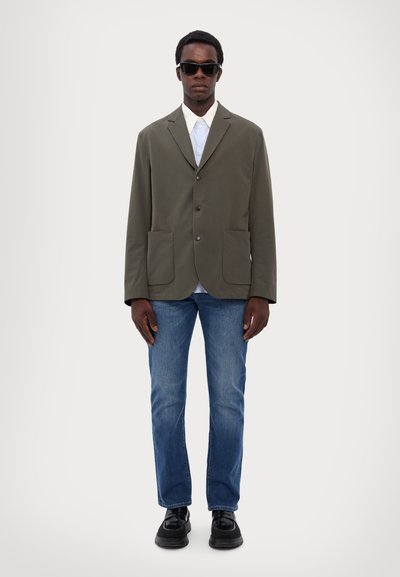 Olive green jacket with a notch lapel, two front pockets, layered over a white shirt with blue stripes, paired with blue jeans and black shoes.