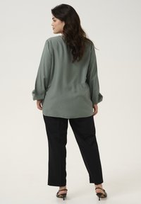 Green textured blouse with long sleeves, featuring a rounded hem, paired with black pants and strappy heel sandals.