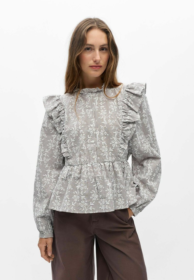 Femme portant un chemisier à manches longues gris à carreaux avec des motifs floraux, des épaules volantées, et un pantalon marron, debout sur un fond blanc uni.