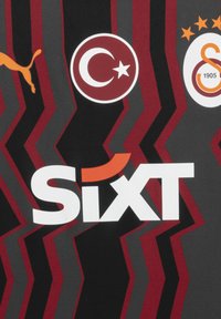 Črn material z rdečim in temno sivim cik-cak vzorcem; vsebuje logotipe: Puma (oranžna), turška zastava in Galatasaray z nalepko "Sixt" v krepki beli in oranžni barvi.