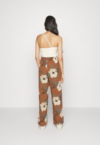 Frau, die mit dem Rücken zur Kamera steht, trägt ein weißes einseitiges Top und braune, blumige Hose mit weitem Bein sowie weiße Plateauschuhe.