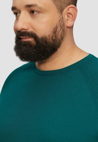 T-shirt en tricot teal à col rond présentant une finition texturée, un tissu lisse et une couture contrastée le long des coutures d'épaule.