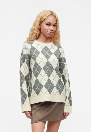 Vrouw met lang krullend blond haar, gekleed in een crèmekleurige en grijze ruiten trui en een bruine minirok, staand tegen een witte achtergrond.