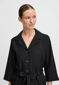 Čierne šaty s gombíkmi, s golierom, trojštvrťovými rukávmi a viazaným pásom, vyrobené z hladkého materiálu s lesklými modrými gombíkmi.