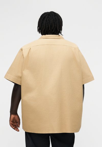 Žmogus su tamsia oda ir trumpais dredais, dėvintis laisvo kirpimo smėlio spalvos trumpomis rankovėmis marškinius, rodomas iš nugaros priešais vientisą foną.