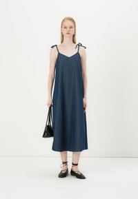 Denim ilgio suknelė su plonais petnešomis ir raištelių akcentais, turinti V formos iškirptę ir laisvą siluetą. Modelis laiko juodą rankinę.