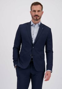 Homme avec des cheveux bruns courts et une barbe portant un costume bleu marine et une chemise habillée blanche à motif devant un fond clair.