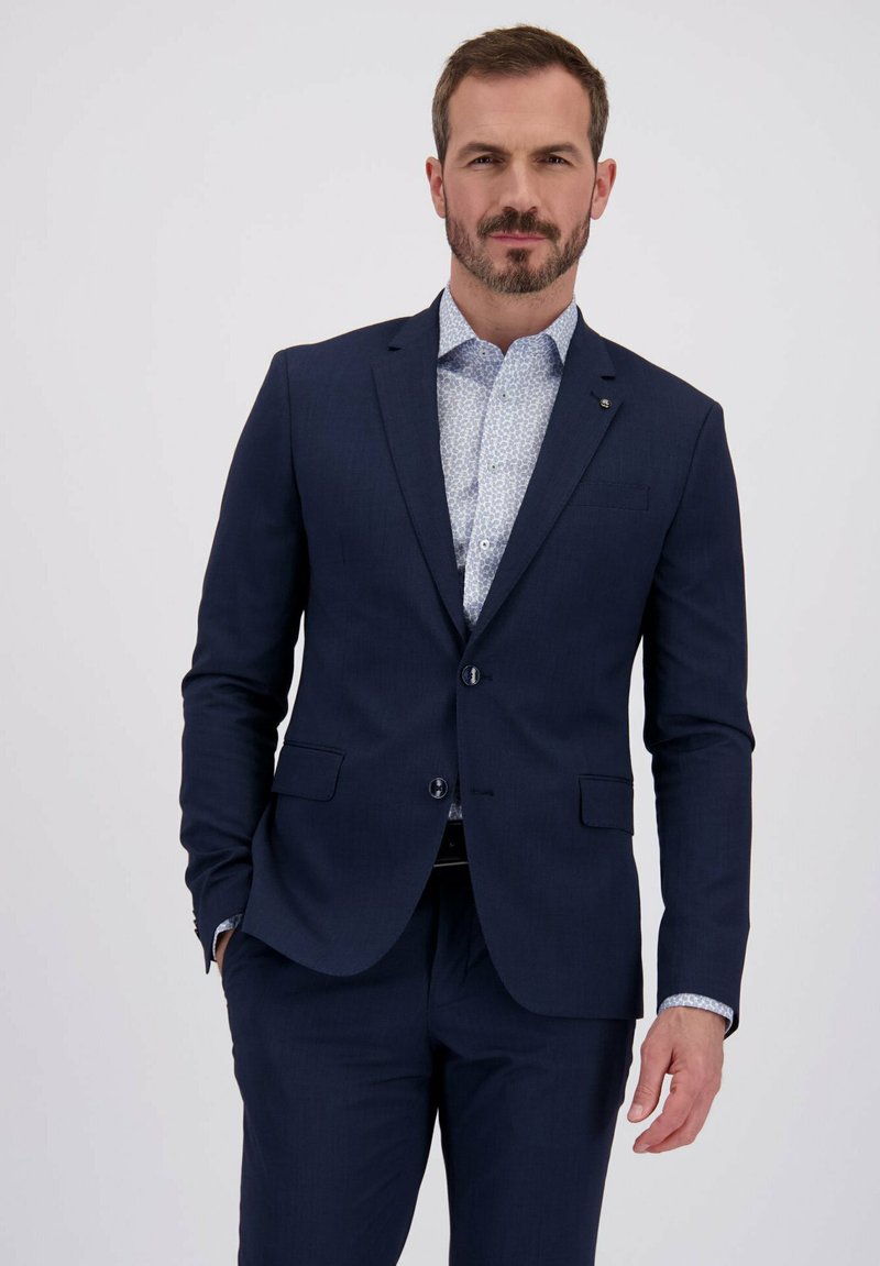 Homme avec des cheveux bruns courts et une barbe portant un costume bleu marine et une chemise habillée blanche à motif devant un fond clair.