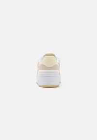Lacoste sneakers med en beige antikk suede-overflate, hvite detaljer og en tykk hvit såle. Har logo på hælen og glatte teksturer.