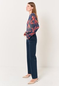 Blusa a maniche lunghe, trasparente, di colore blu navy con motivo floreale rosso; abbinata a pantaloni blu navy e scarpe con tacco nude. Modella mostrata di profilo.
